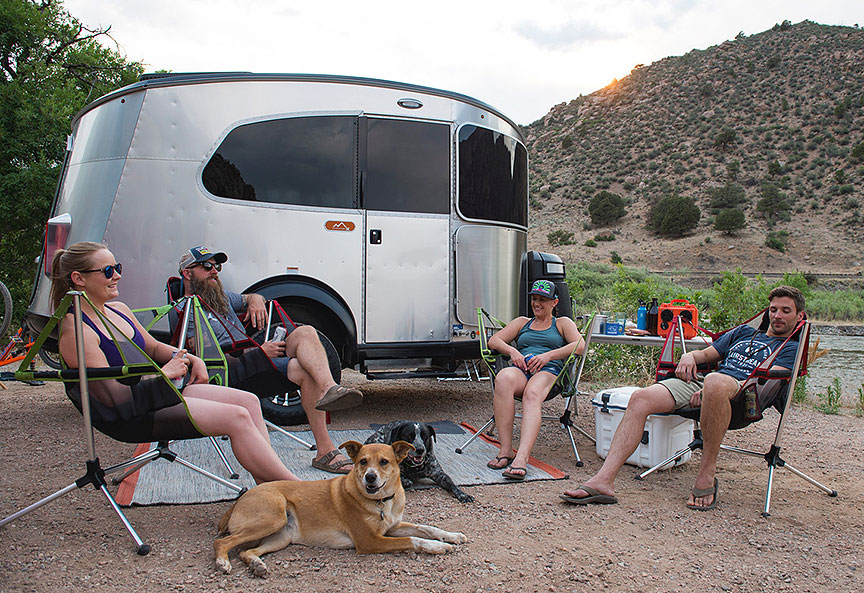 Kicking back outside the Airstream Basecamp. Four people in camp chairs and a dog in front of the Basecamp trailer.
